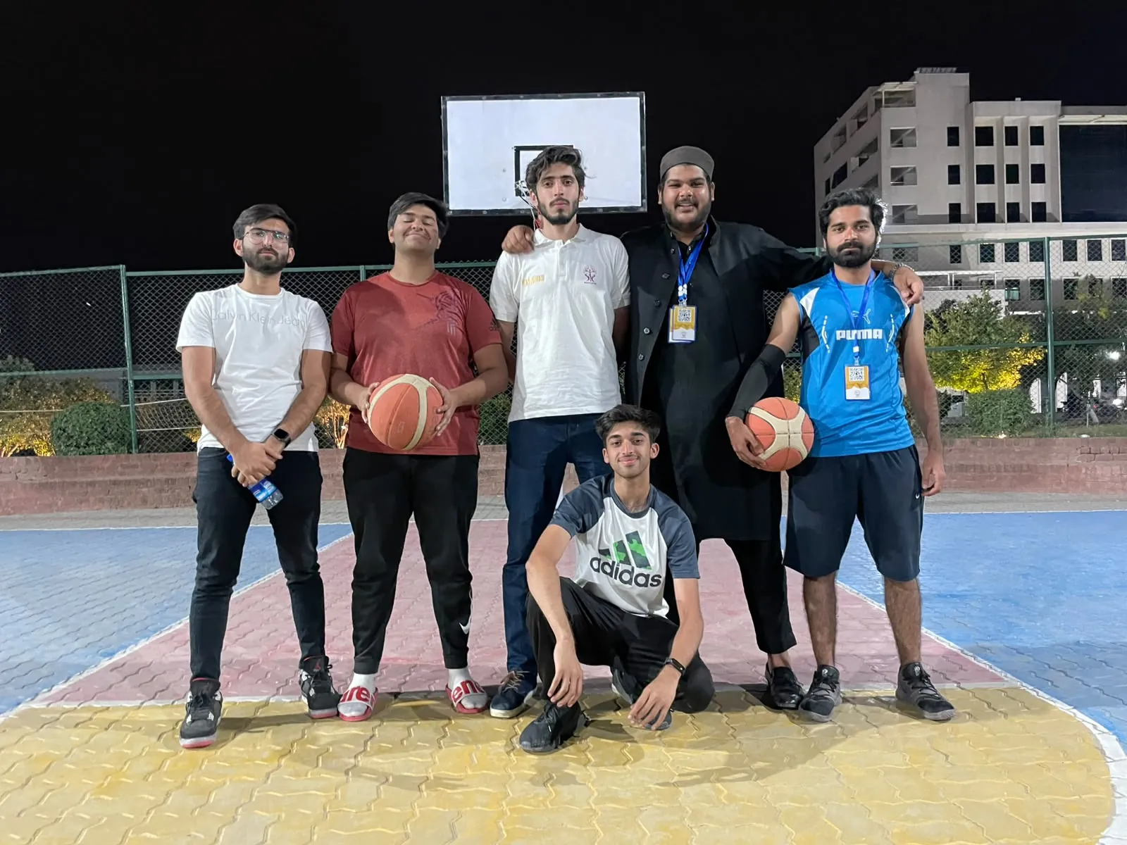 Six players posing under the hoop on an outdoor court at night.
